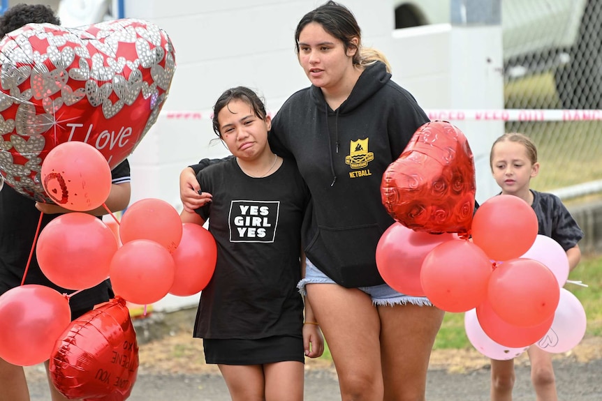 A teenager comforts a young girl crying as they bring red balloons to a blessing service