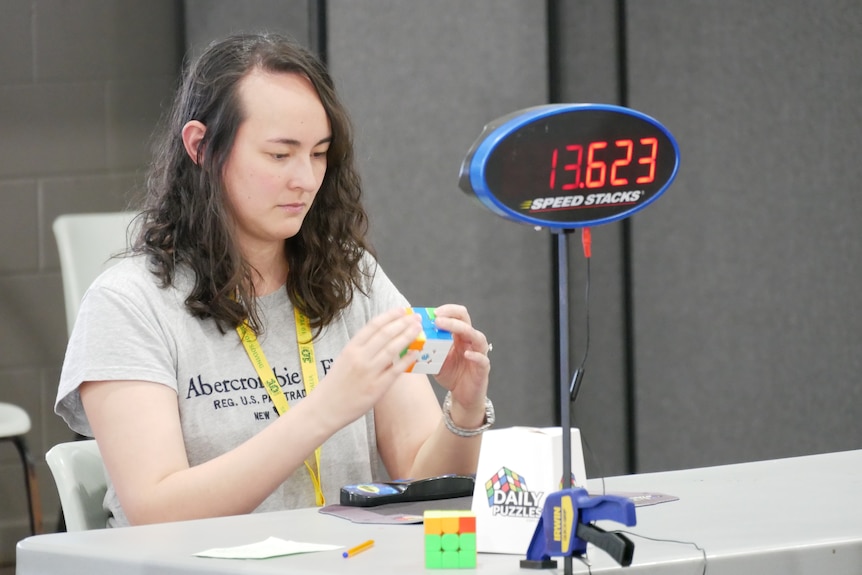 A woman in a grey shirt sits at a table with a timer solving a Rubik's Cube.