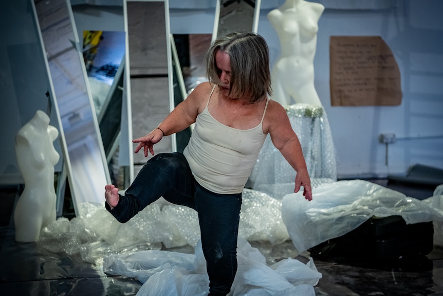 Leisa Prowd lifts one leg in the air during a dance, in front of three mirrors and two mannequins.