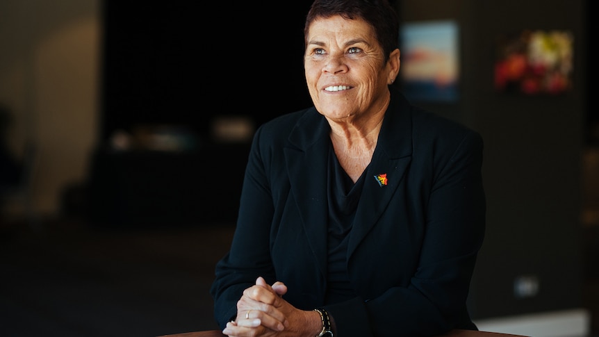 A mature Indigenous woman in a black blouse sits at a table, hands clasped, smiling and looking to the left of the frame.
