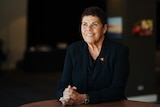 A mature Indigenous woman in a black blouse sits at a table, hands clasped, smiling and looking to the left of the frame.