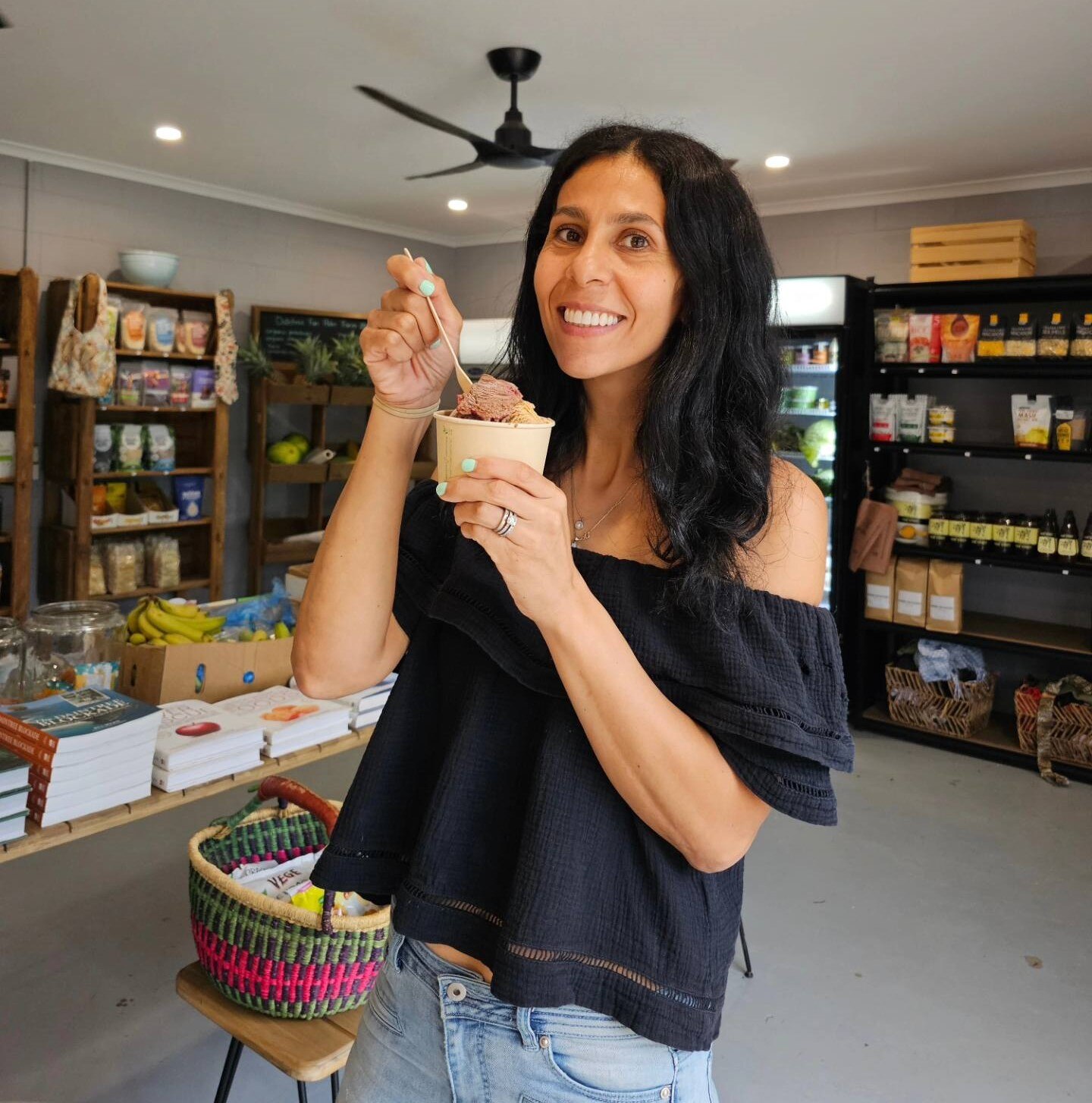 A woman is standing in a store, holding a small cup of ice cream and smiling.