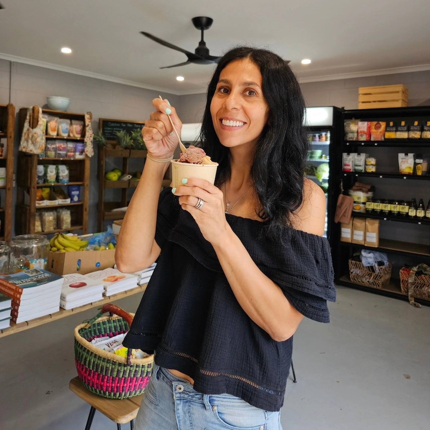 A woman stands in a general store, holding a little cup of ice cream and smiling.