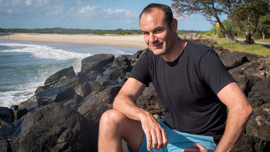 Man sits on a coastal rock wall at East Ballina with beach behind him.