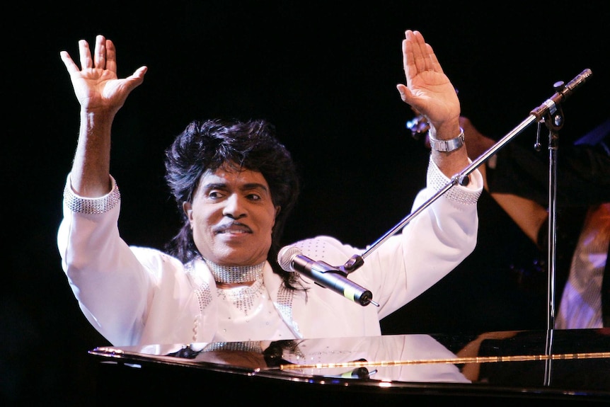Dressed all in white and sitting at his piano, Little Richard smiles and waves to the crowd.