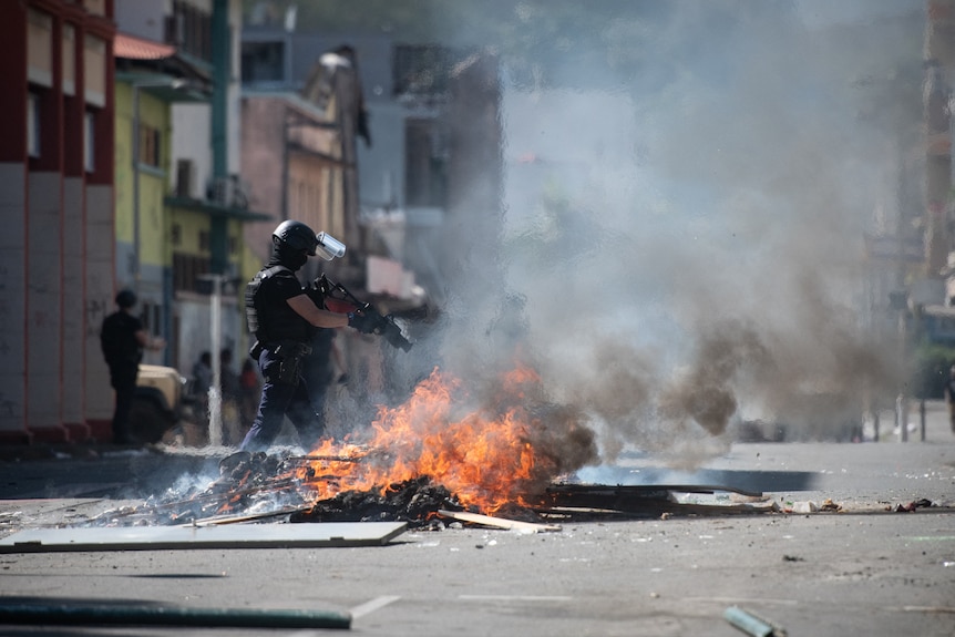 An armoured police officer with a gun stands over a flaming roadblock.