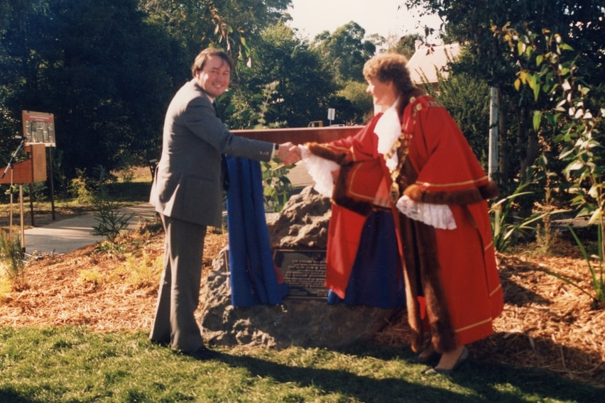 An old colour photograph of a man in a suit smiling and shaking hands with a woman in a mayoral gown.