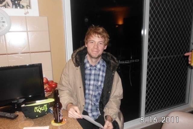 A man wearing a blue chequered shirt sitting at a kitchen table holding a letter, smiling