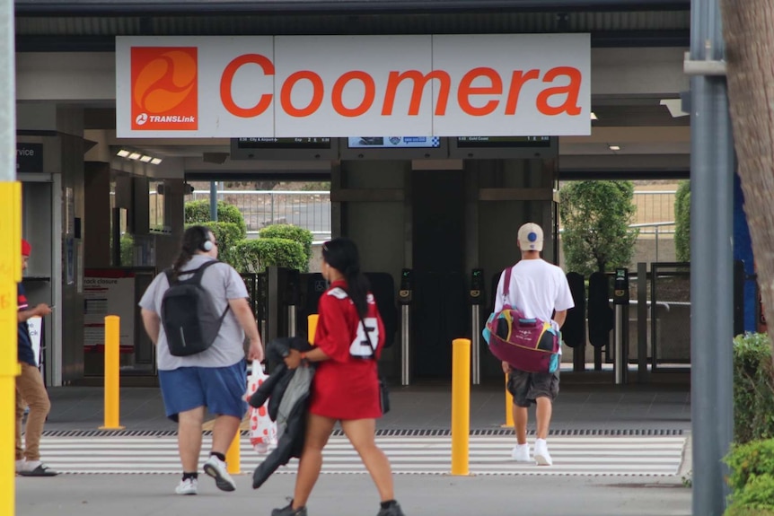 The entrance to Coomera train station with an orange sign and people walking through the entrance.