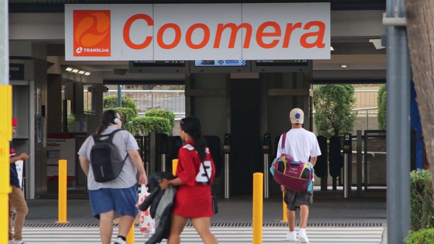 The entrance to Coomera train station with an orange sign and people walking through the entrance.