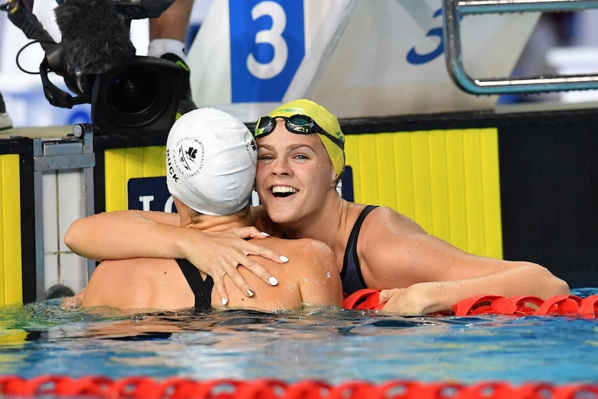 Two female swimmers wearing caps and goggles on their head hug post-race