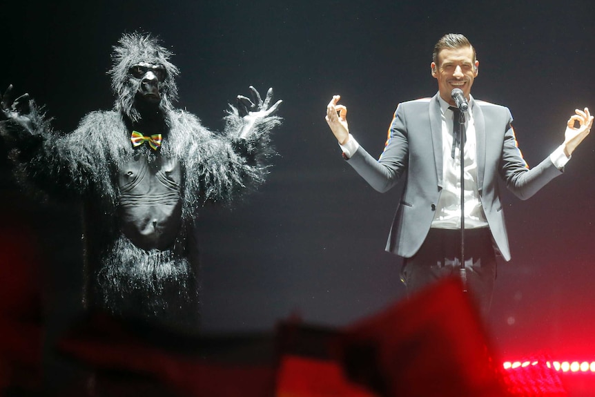 Francesco Gabanni wears a black suit with rainbow trim accompanied by a man in a full gorilla suit on stage
