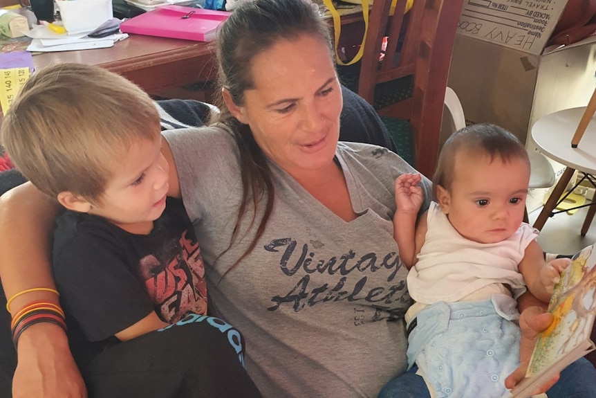 Two children sit on their mother's lap while she reads them a children's book.