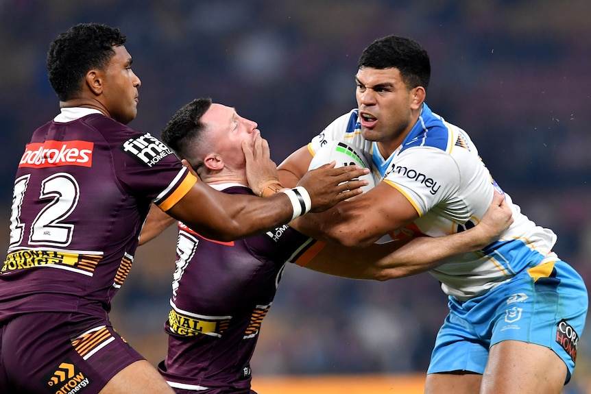 A Gold Coast Titans NRL player holds the ball as he attempts to fend off two Brisbane Broncos opponents.