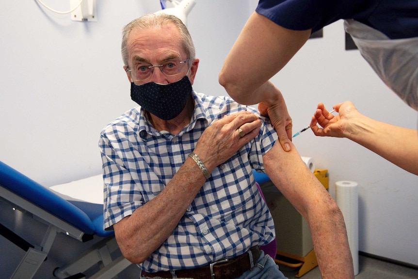 An elderly man in a blue check shirt wearing a mask receives a vaccination.