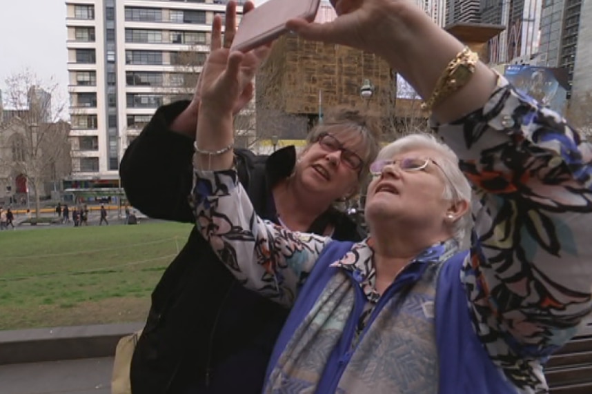 Two women standing outside look up into a smart phone to take a selfie.