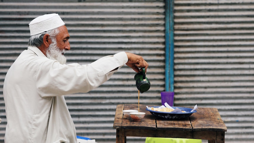An old man pouring tea into his cup sitting at a table.