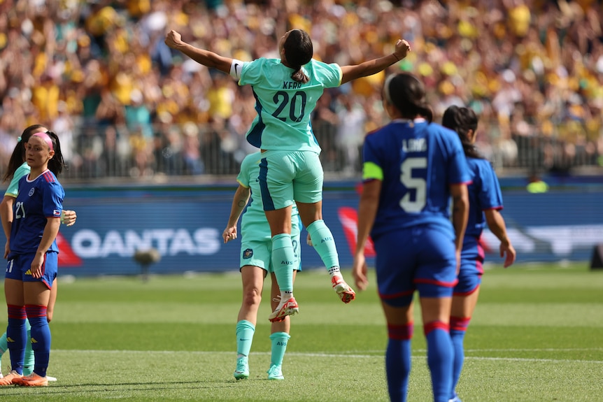 Matildas star Sam Kerr throws her arms wide as she leaps in the air in celebration after scoring in an international.