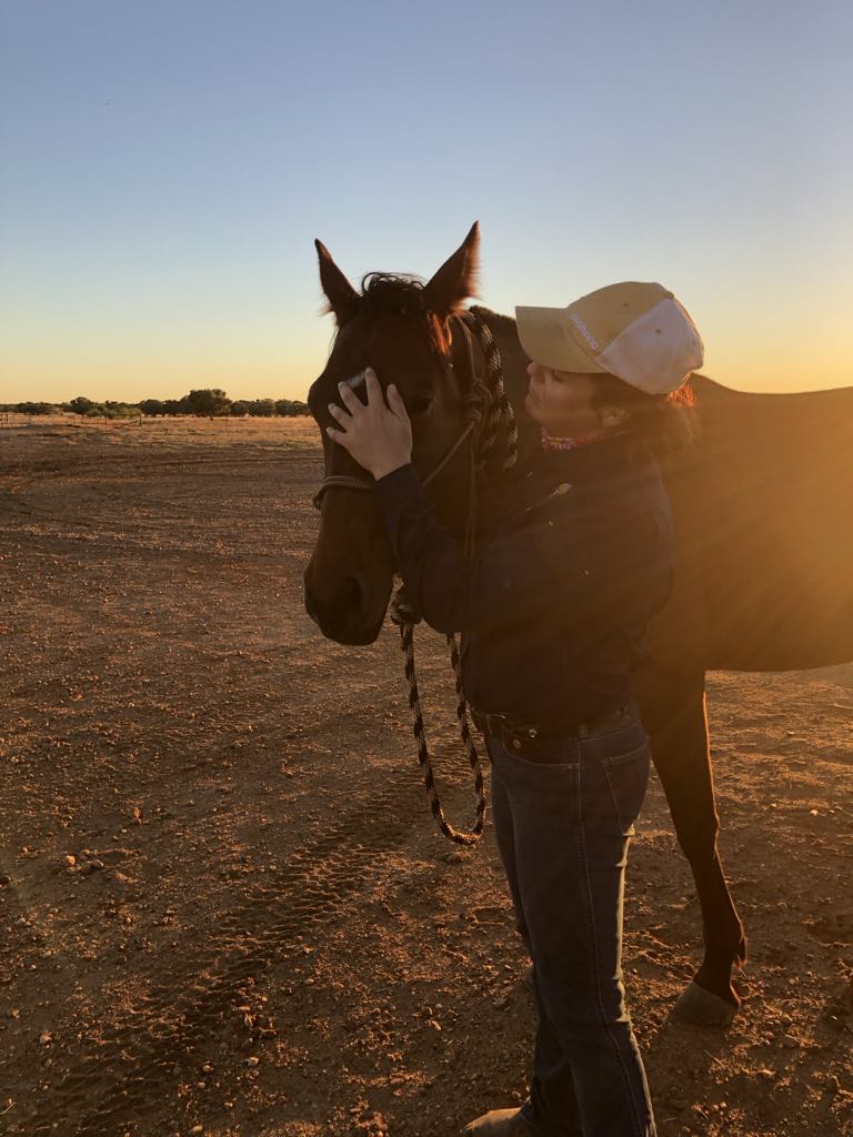 A woman wearing a cap pats her horse as the sun sets over burnt orange dirt