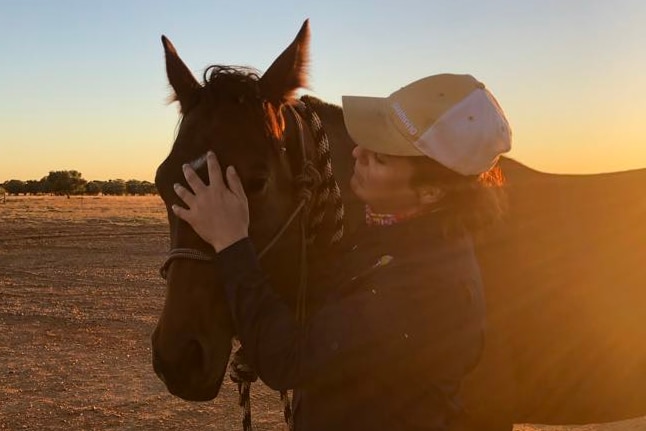 A woman wearing a cap pats her horse as the sun sets over burnt orange dirt
