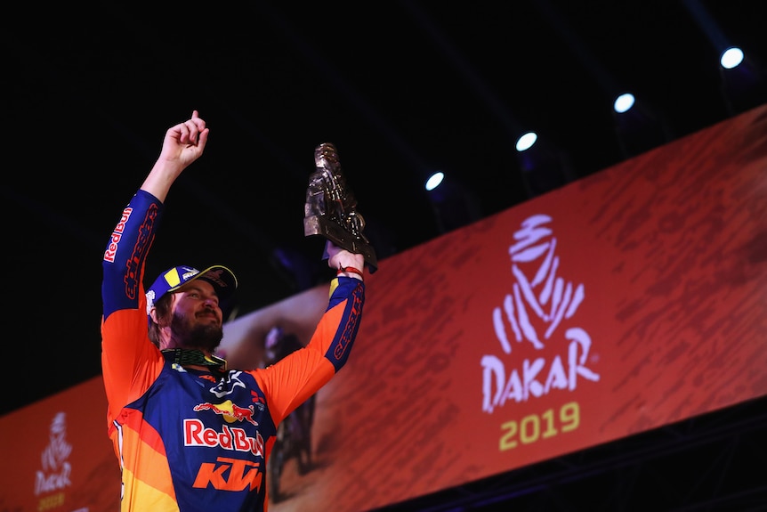 A motorcycle racer holds a trophy one hand with his other hand pointing skywards near a sign saying 'Dakar 2019''.
