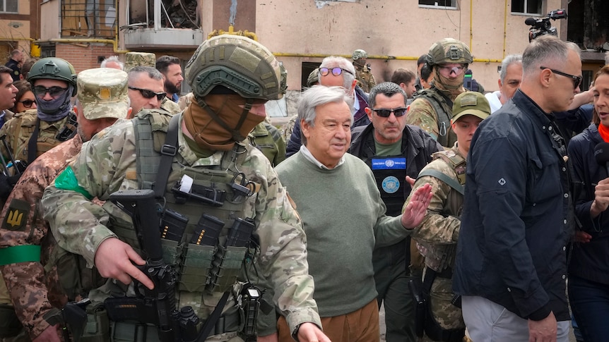 Man in green sweater surrounded by armed soldiers.