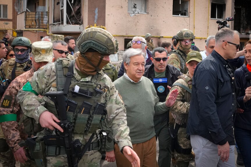 Man in green sweater surrounded by armed soldiers.