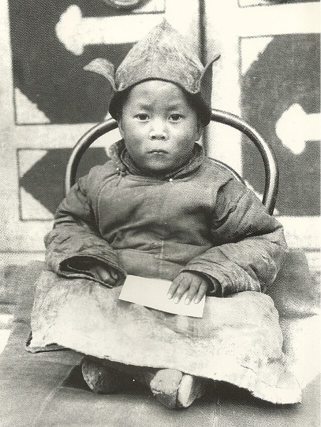 A solemn-faced young child sits on a wooden chair with his legs crossed. He wears a hat