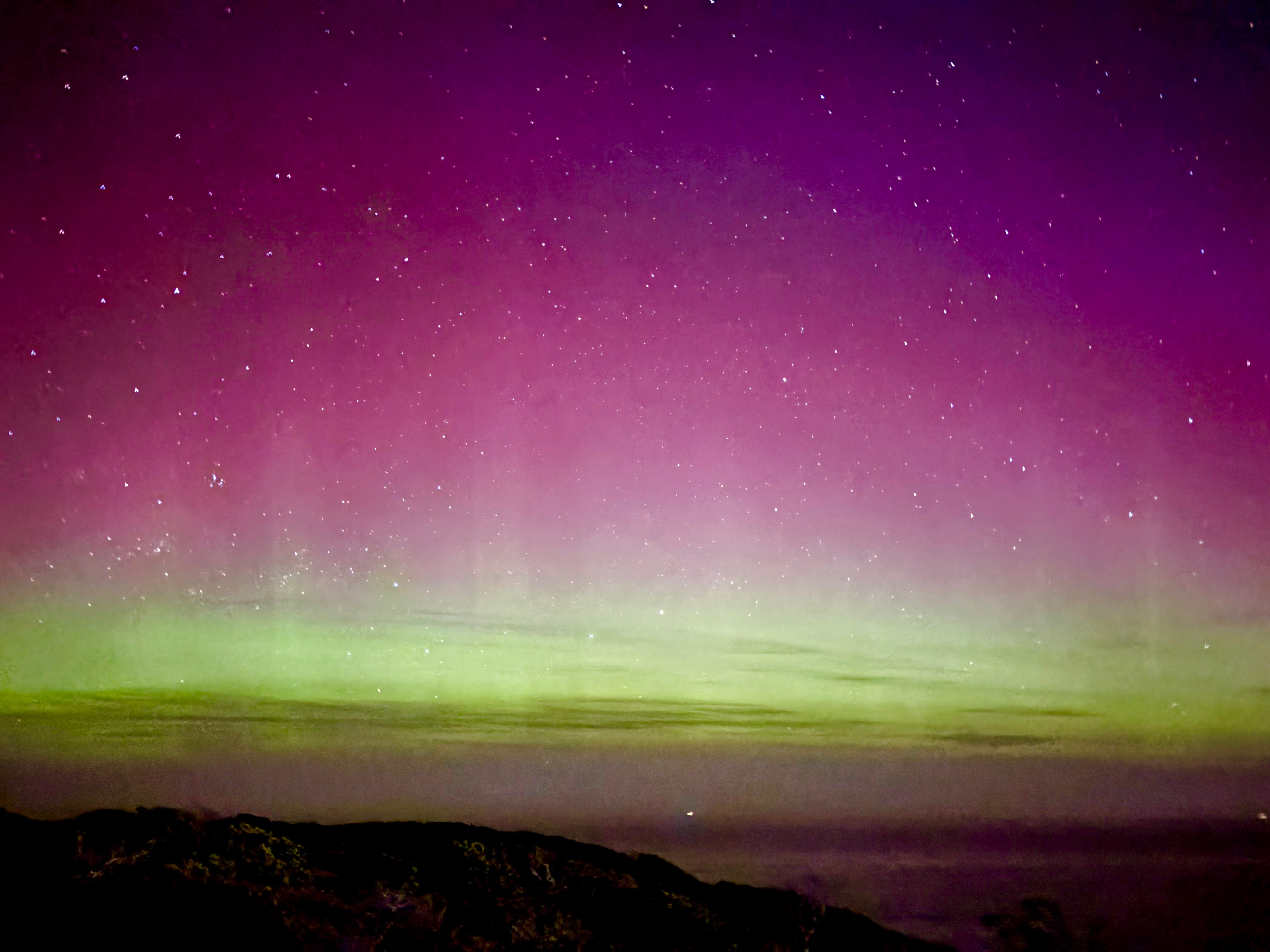 Uma faixa de luz verde-amarela no horizonte com tons profundos de rosa e roxo acima, com centenas de estrelas no céu.