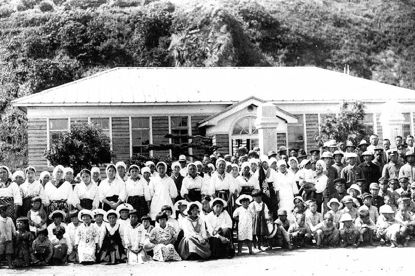An old black and white photograph of a big group of Japanese people gathered in front of a small house