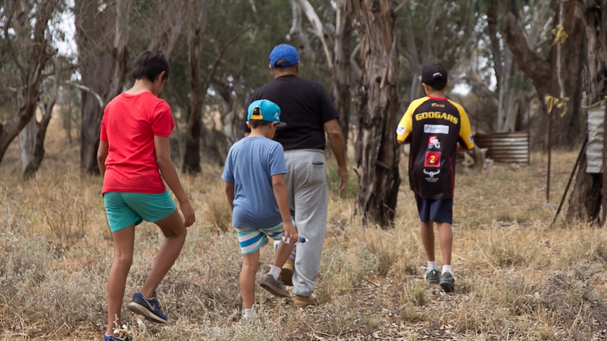 A man and three boys walking through bushland with some old corrugated iron the background