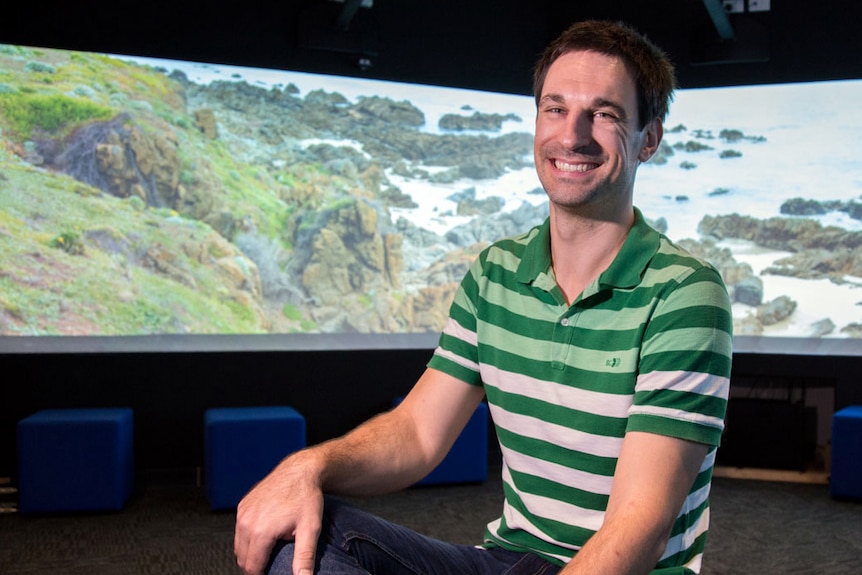 Dr Raimondo sits in an exhibition space with a graphic of a coastline behind him.