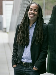 Jonathan W. Gray with long locs, smiling, wearing a black suit and blue shirt, no tie