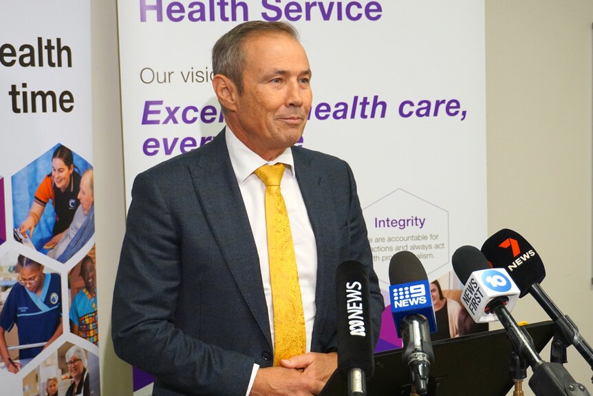 Premier Roger Cook in a suit jacket and yellow tie, standing behind a row of microphones.