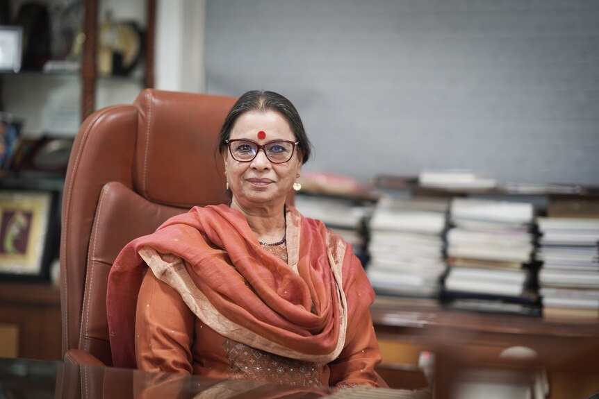 A close up of a women wearing an orange sari sitting at her desk in an orange chair. A stack of books sit in a pile nearby.
