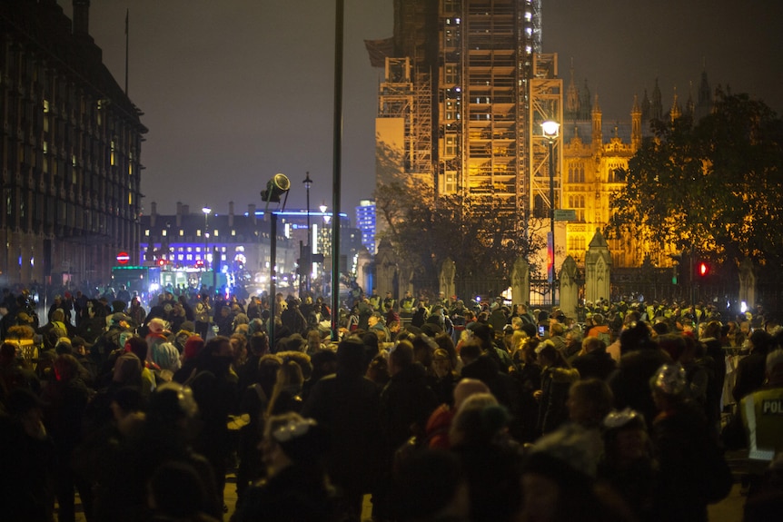 A huge crowd gathers in a public square, looking towards an illuminated building partly that is covered by scaffolding.
