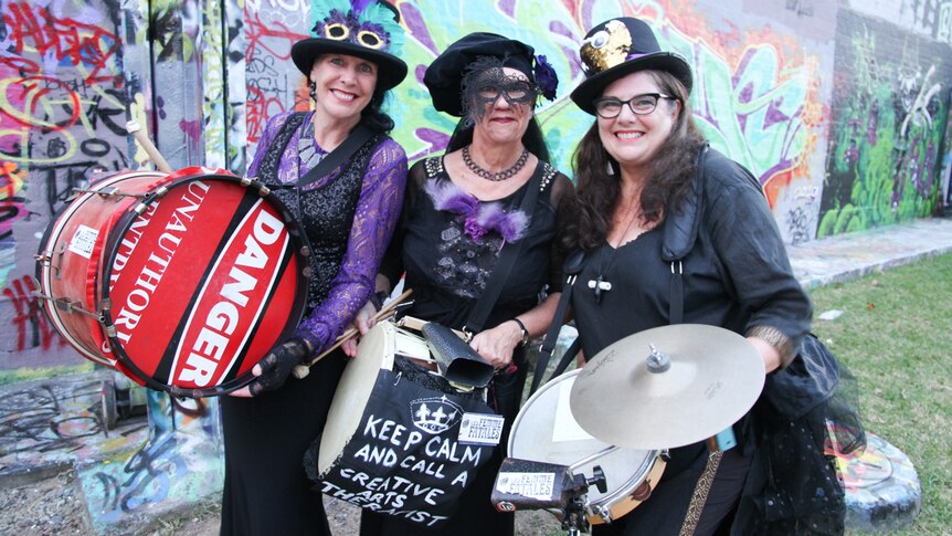 Three women, dressed in black and purple and carrying musical instruments, smile to camera