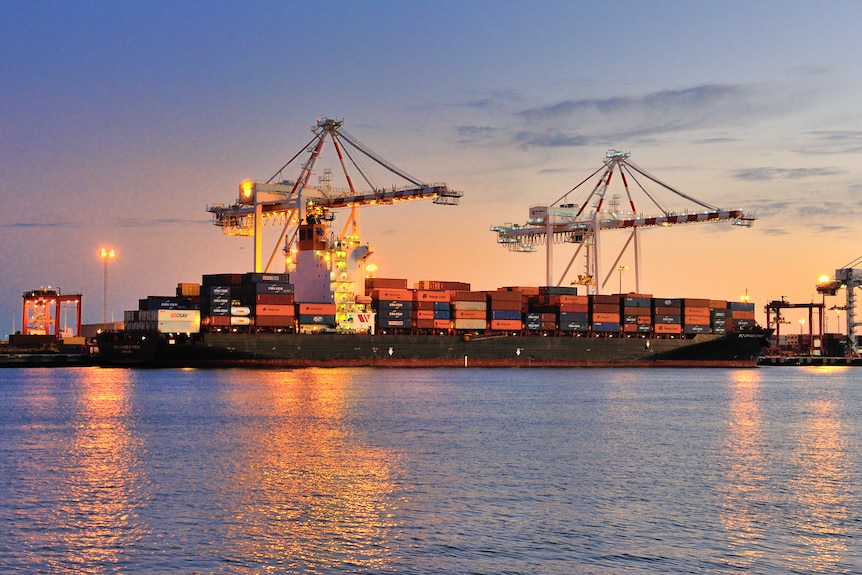 A large container ship alongside a wharf at sunset, with an evening glow in the background and lights reflecting on the water.