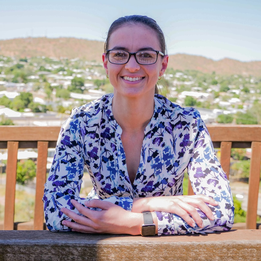 A woman with dark hair pulled back, wearing glasses and a blue and white shirt, sitting at a lookout in Mount Isa.