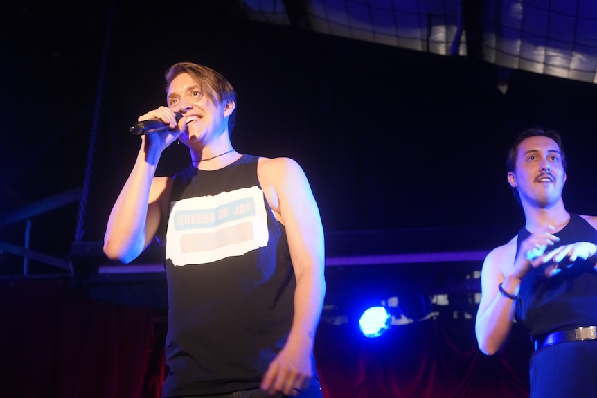 Person performing on stage, holding a microphone and wearing a black tank top. Another person is by their side.