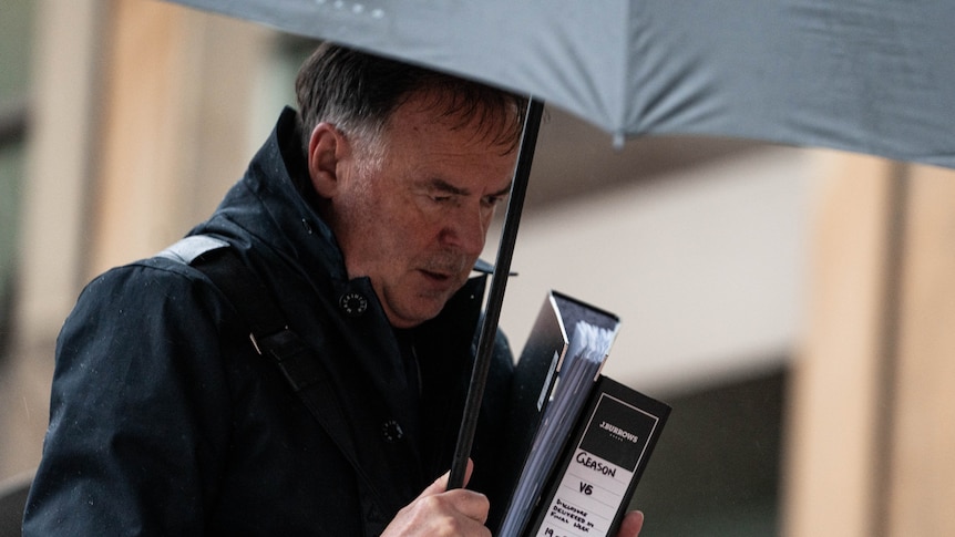 Man walking into court holding documents and covering his face with an umbrella.