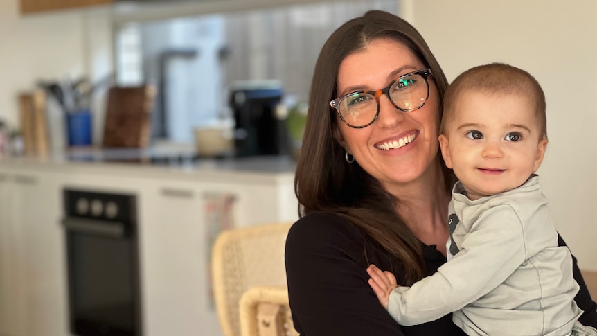 A smiling mother sits in her kitchen and holds her baby.