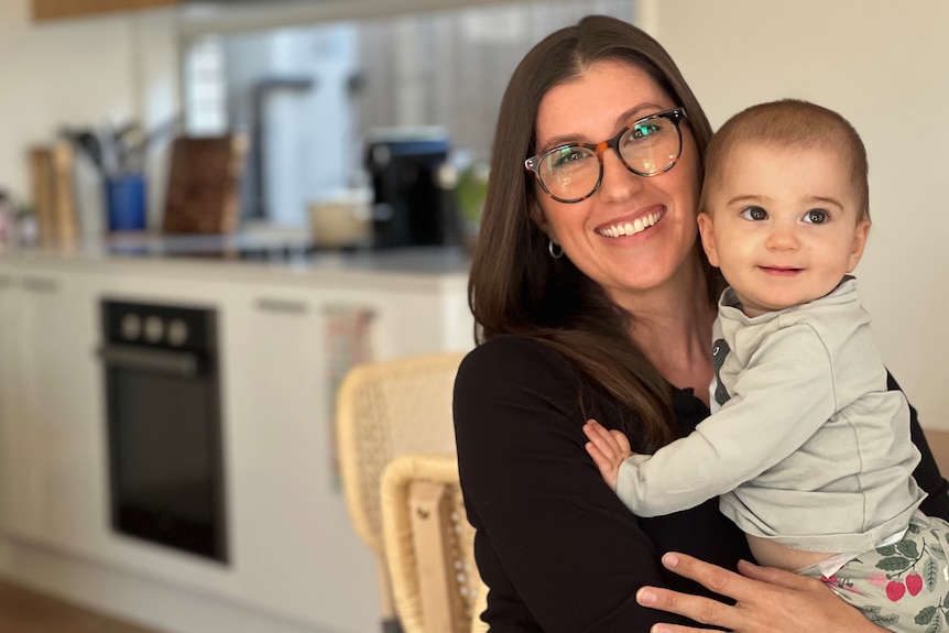 A smiling mother sits in her kitchen and holds her baby.