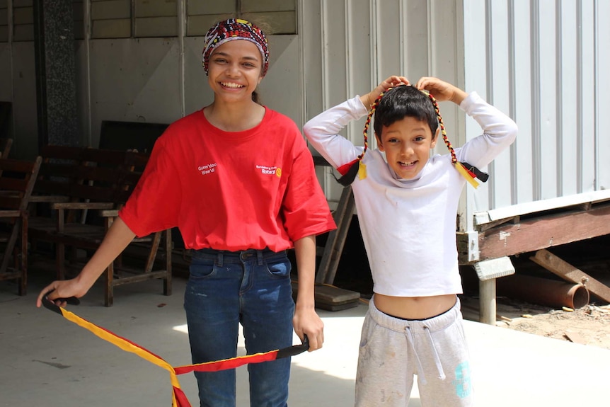 A girl wearing a headscarf smiles and stands next to a younger boy holding up a plait in red, white and black colours.