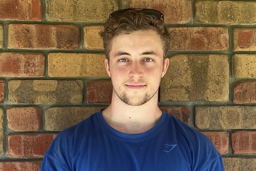 a young caucasian man in a blue shirt standing in front of a brick wall