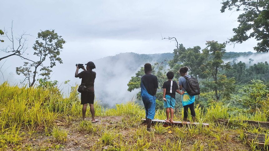 Three older kids and one younger look out over a lush valley where fog is lifting. An older one is holding a DSLR camera.
