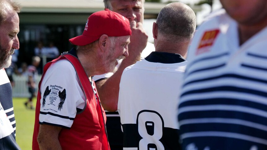 An older man with a red cap surrounded by other men in rugby union jerseys.