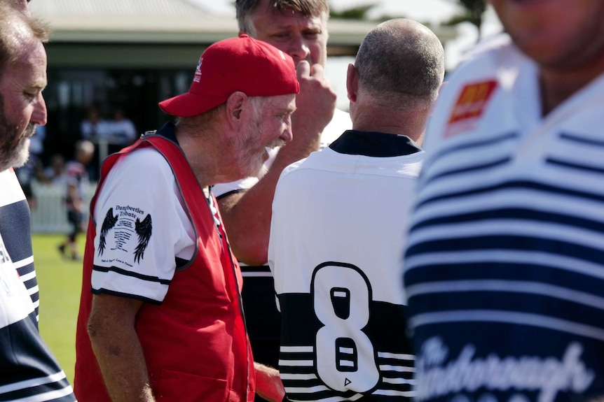 An older man with a red cap surrounded by other men in rugby union jerseys.