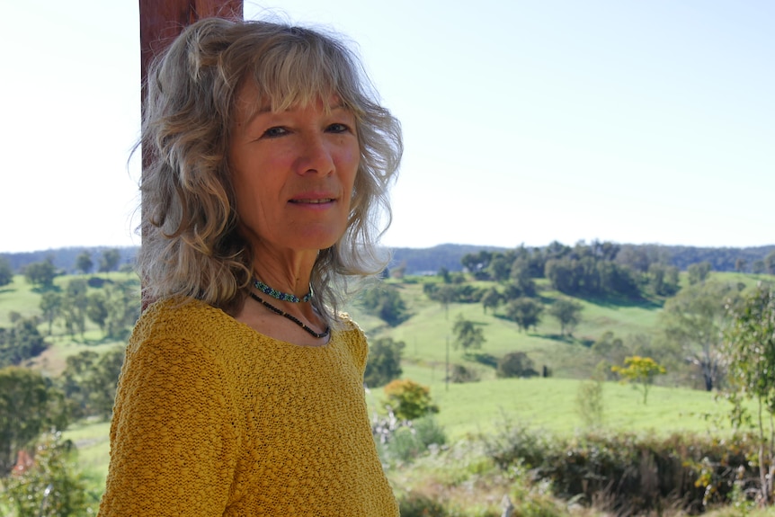 Woman with long grey hair wearing yellow sweater. backdrop of rolling green hills