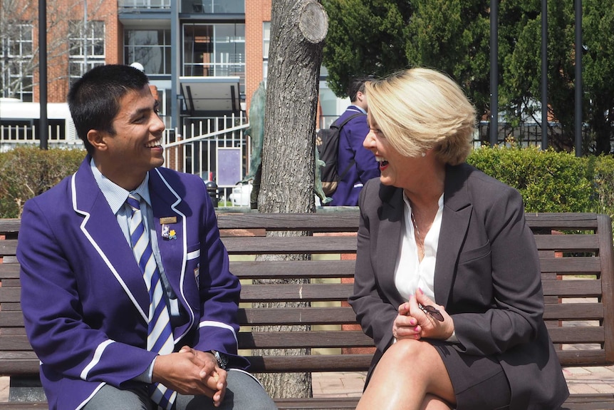A teenage boy in a school uniform sits on a bench with the school's counsellor